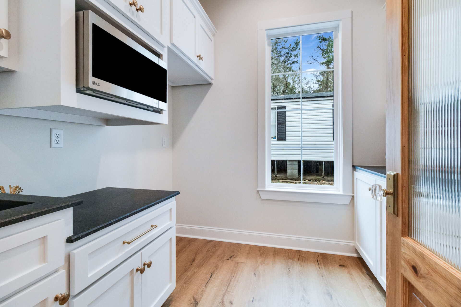 A small, well-lit kitchen with white cabinets and drawers, a black countertop, and a built-in microwave. A window provides natural light and a view of an exterior Trahan structure. There is a wooden door with a frosted glass panel to the right.