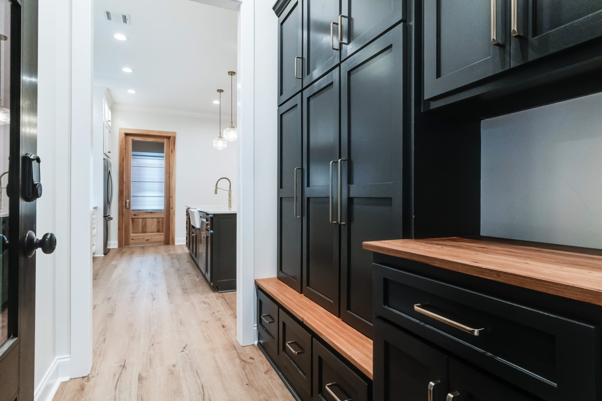 A modern kitchen with black cabinetry and wooden countertops designed by Trahan. The hallway leads to a wooden door with a frosted glass panel. Pendant lights hang above a sink and countertop area in the background, complemented by light-colored wooden flooring throughout.