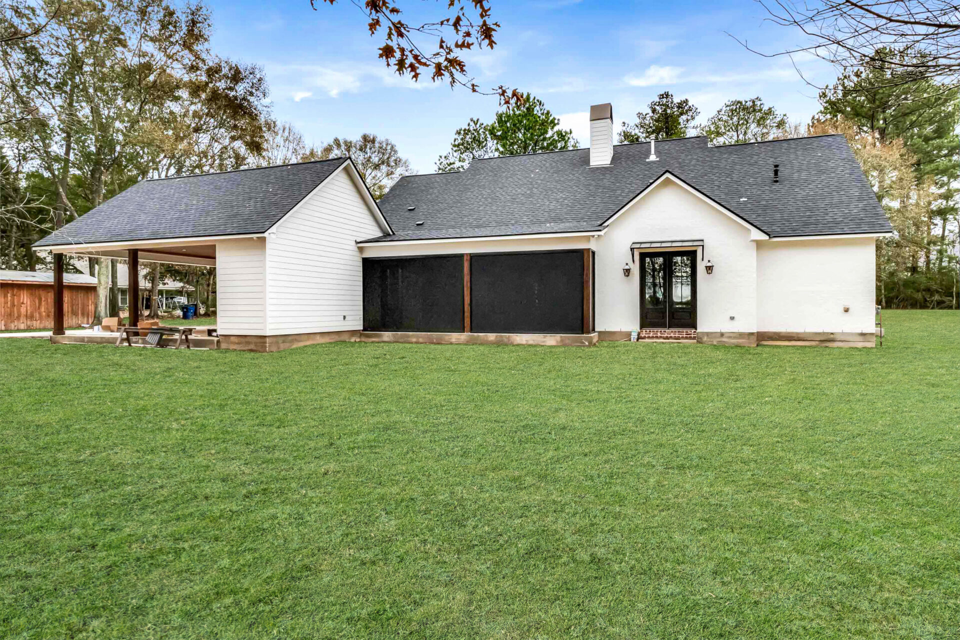 A single-story Trahan house with white exterior walls, black shutters, and a black roof. The house features a screened-in porch, a chimney, and a patio area with a picnic table covered by an extended roof. The property is surrounded by grass and trees.