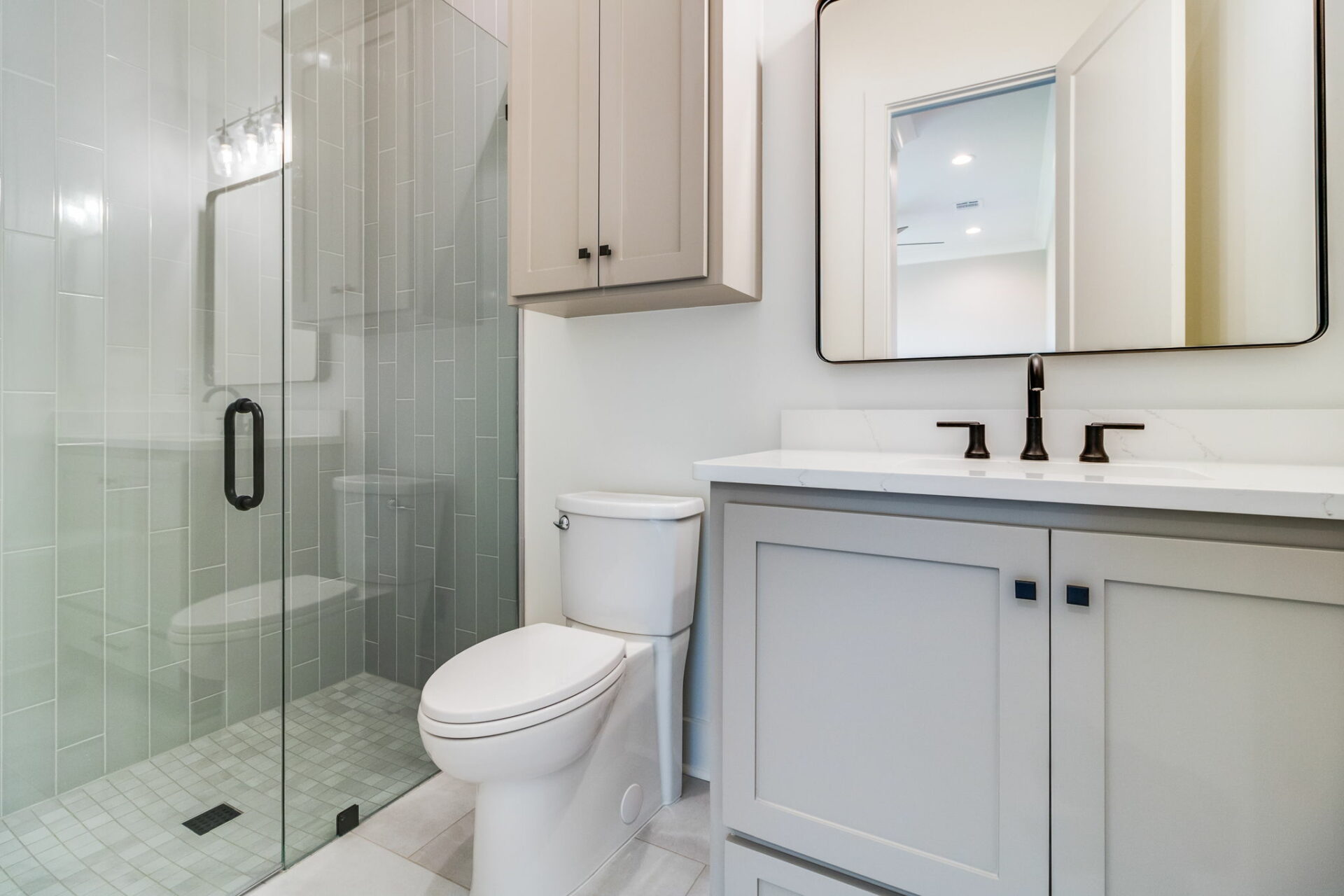 A modern bathroom featuring a glass-enclosed shower, grey-tiled walls, a white Waltman toilet, and a grey vanity with a white countertop. The vanity boasts a large rectangular mirror and sleek black faucets, complemented by cabinets above and below for ample storage.