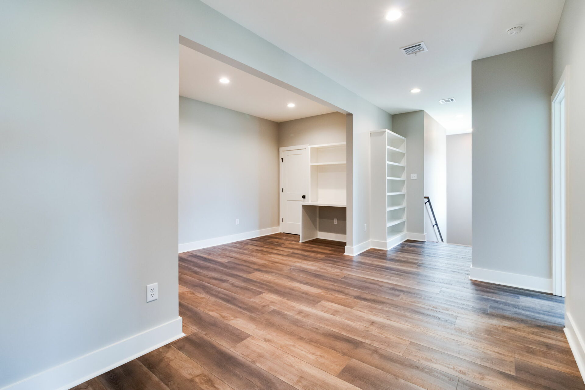 A room with light gray walls and dark wood flooring. The open space features a built-in white desk and shelves in a niche. Recessed lighting adorns the ceiling, highlighting the sophisticated design. A doorway, possibly leading to another section of the Waltman home, is visible in the background.