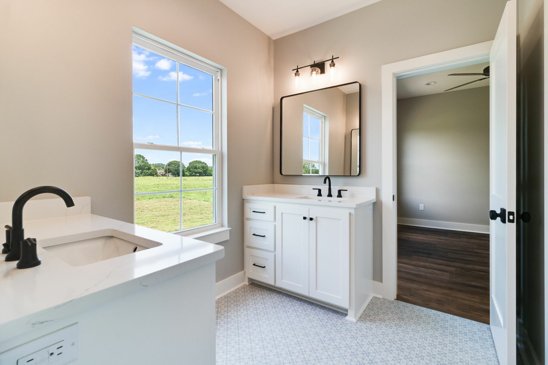 A bright bathroom in the Waltman residence features a large window with a scenic outdoor view, a rectangular mirror above a white vanity with black fixtures, a white countertop with a sink, and light grey walls. The adjoining room has wood flooring and a ceiling fan.