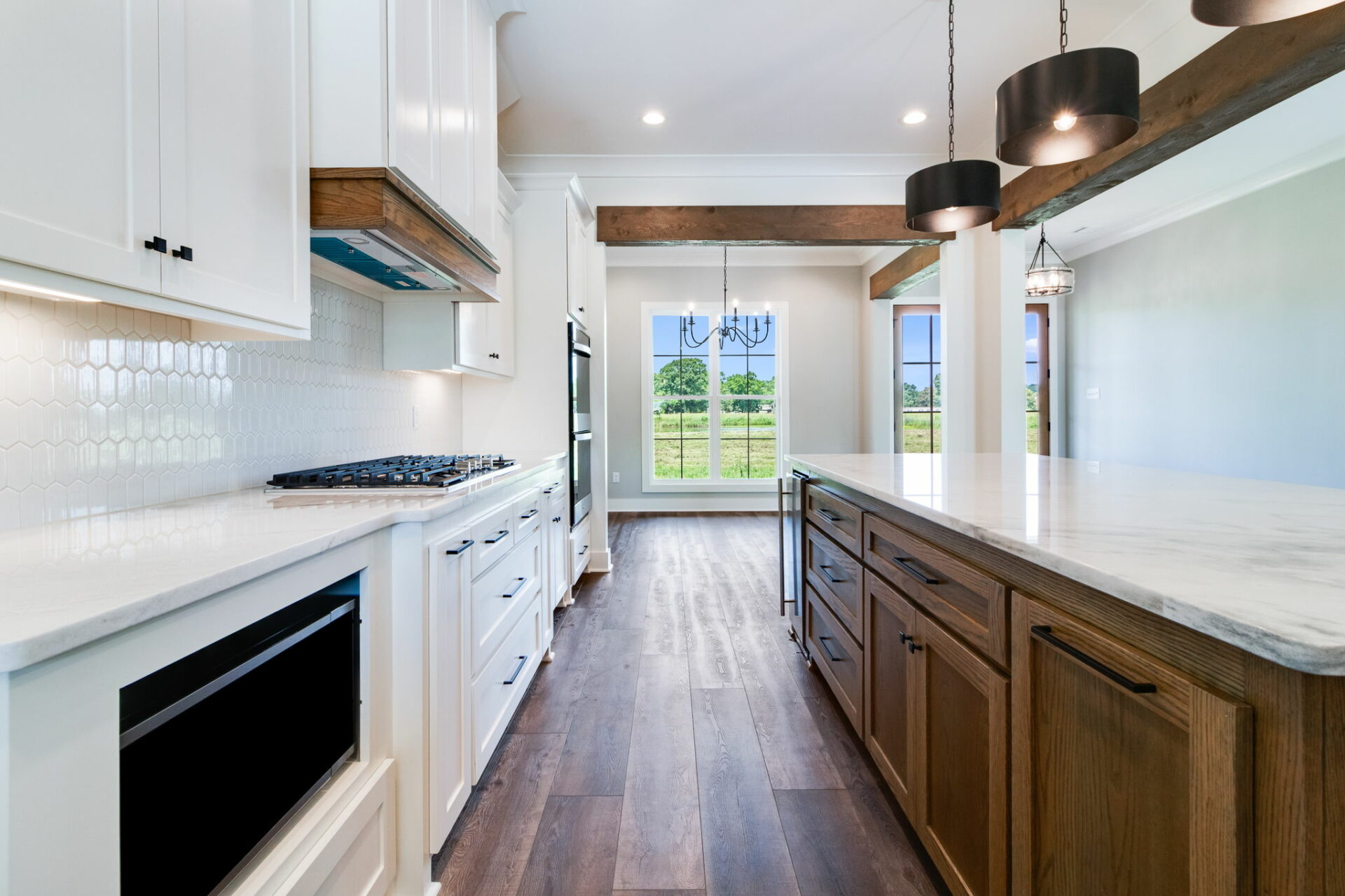 A modern Waltman kitchen with white cabinets, a white backsplash, and stainless steel appliances on the left side. To the right, there is a large island with a marble countertop and brown cabinets. Two pendant lights hang over the island, and large windows are visible at the far end.