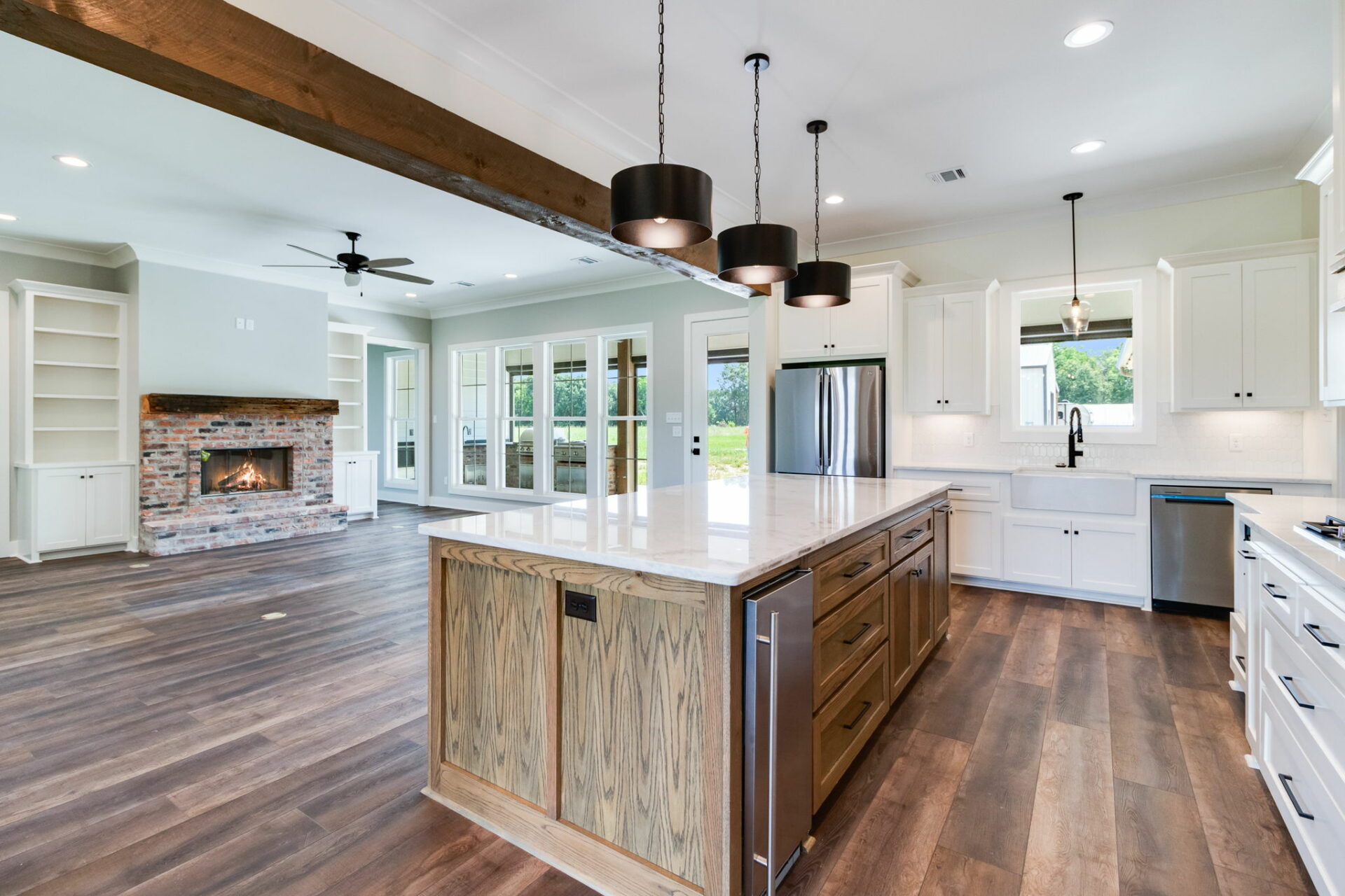 A spacious kitchen and living area featuring wooden floors and white cabinets. A large central island with a wooden base and marble countertop holds three pendant lights above. The background shows a brick fireplace, a wall of windows, and signature Waltman design elements.