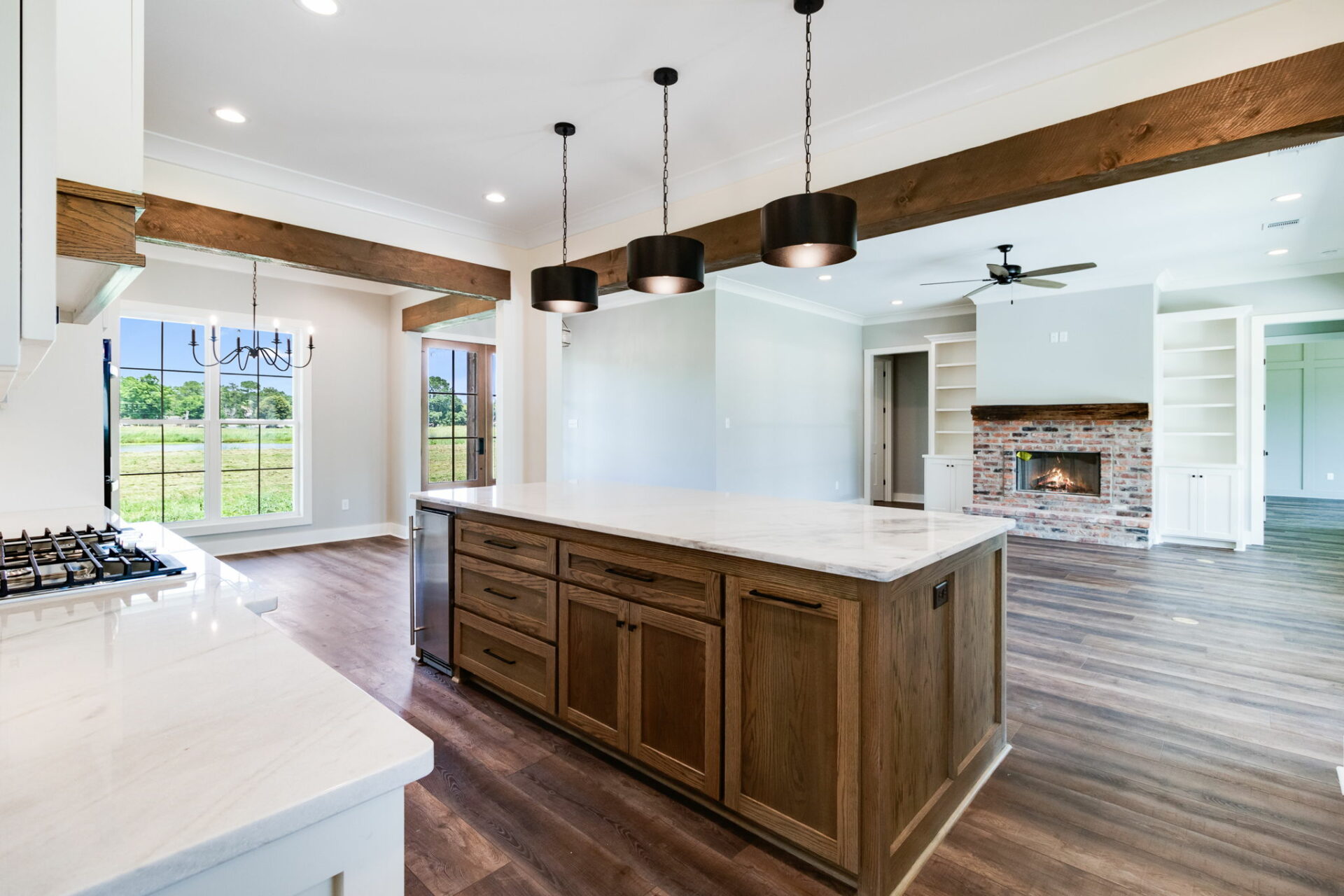 A modern kitchen and living area with hardwood floors. The Waltman kitchen features a large island with a marble countertop and three hanging pendant lights. The living area has a brick fireplace, built-in shelves, and a ceiling fan. Large windows provide natural light.