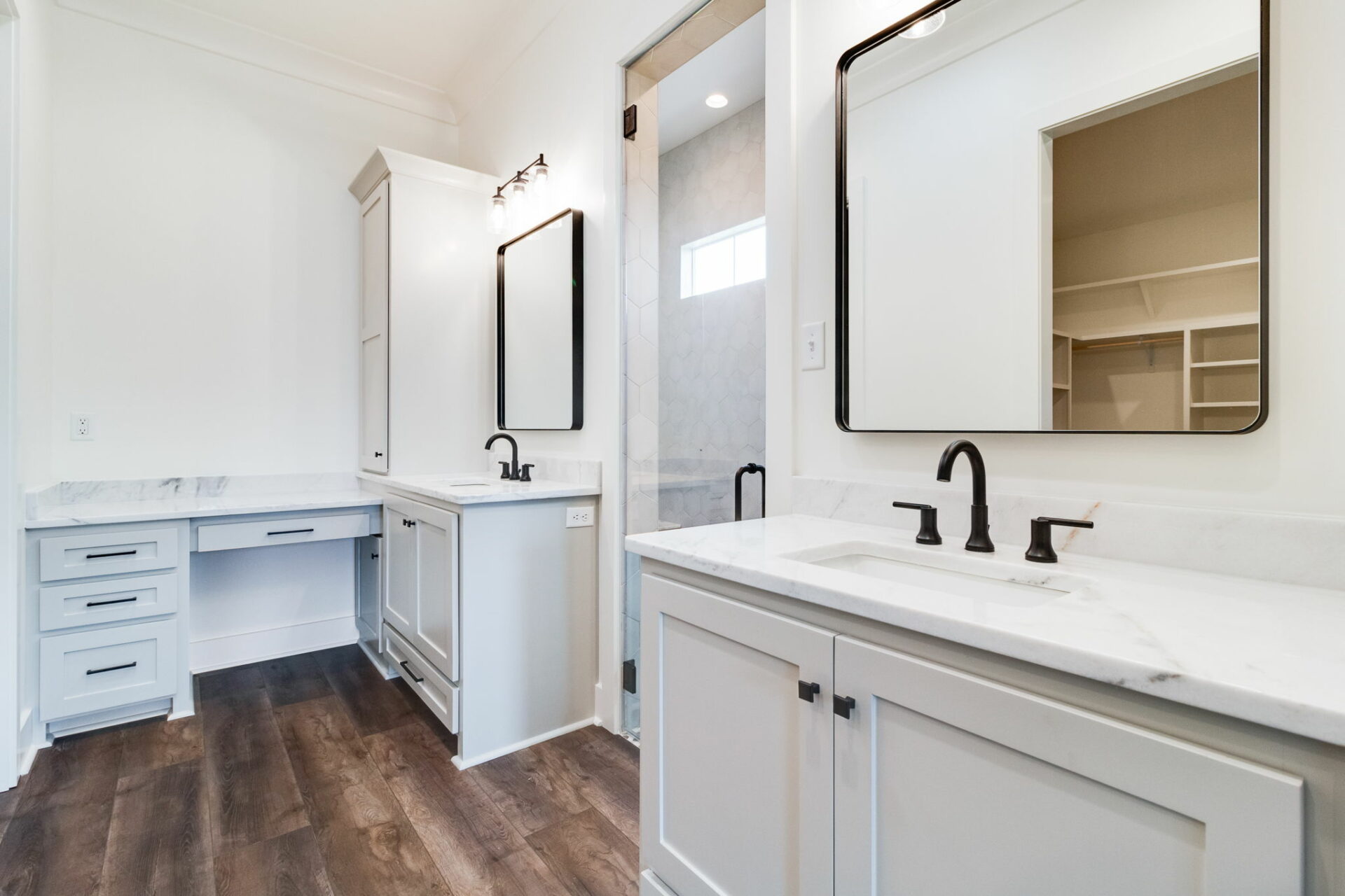 A modern Waltman bathroom with white cabinetry and light marble countertops. It features two rectangular mirrors, black faucets, and a built-in vanity area. The space has wood flooring and neutral-colored walls, while a glimpse of a walk-in shower is visible in the background.