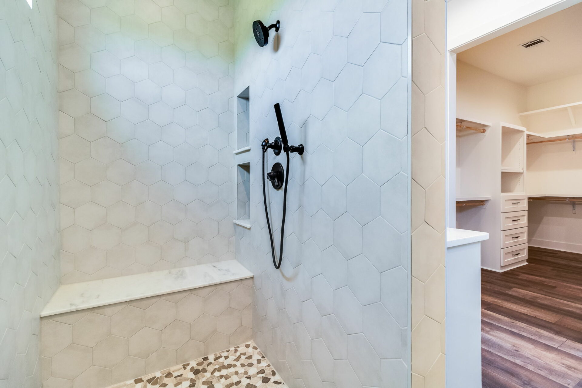 A modern Waltman shower with hexagonal white tiles on the walls and terrazzo flooring. The shower features a black handheld and mounted showerhead, and built-in niches. Adjacent to the shower is a walk-in closet with wooden flooring and white built-in shelves and drawers.