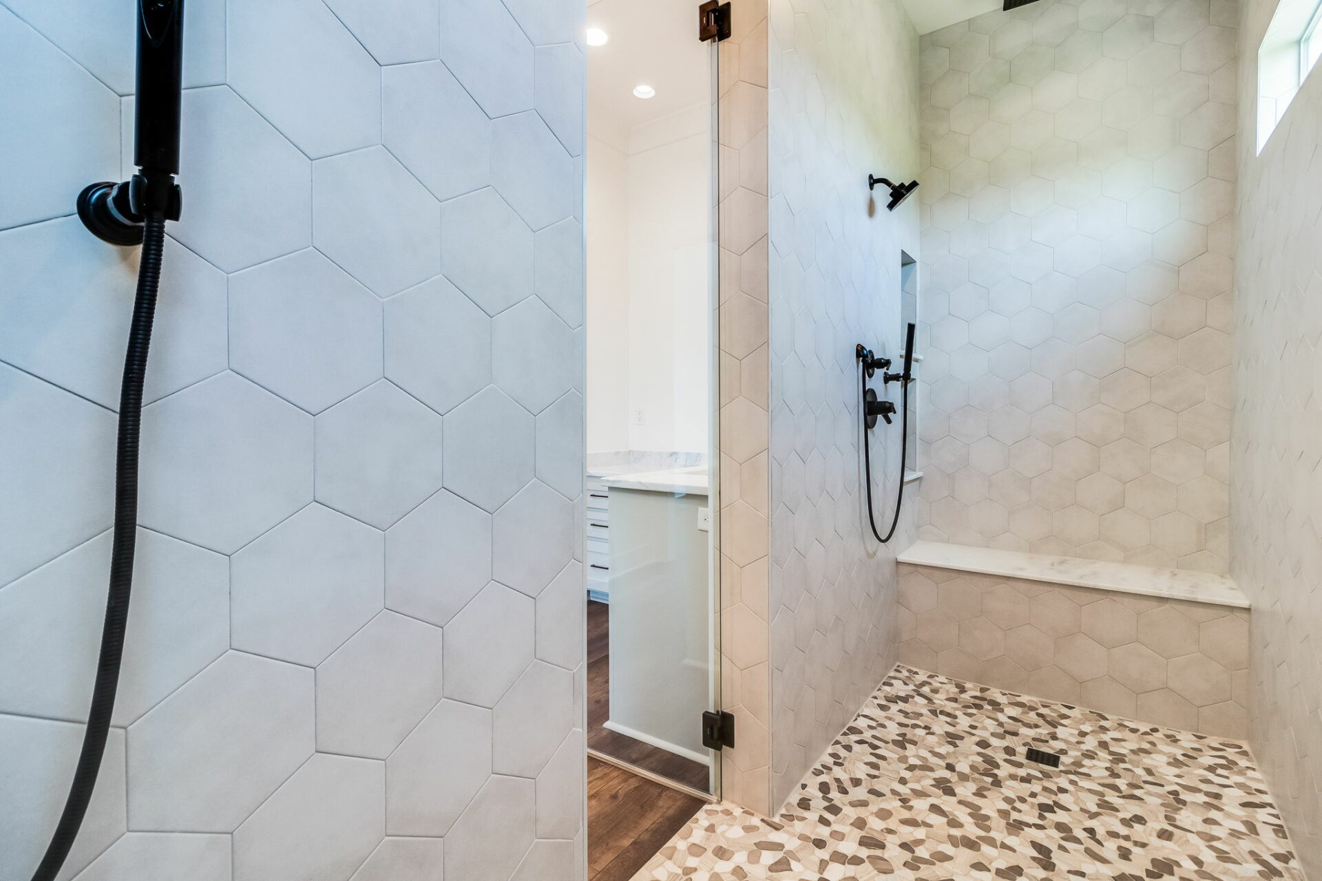 This image shows a modern walk-in shower in the Waltman design, featuring large hexagonal tiles on the wall and small pebble-like tiles on the floor. The shower includes black fixtures, a glass door, and a built-in bench along the back wall.