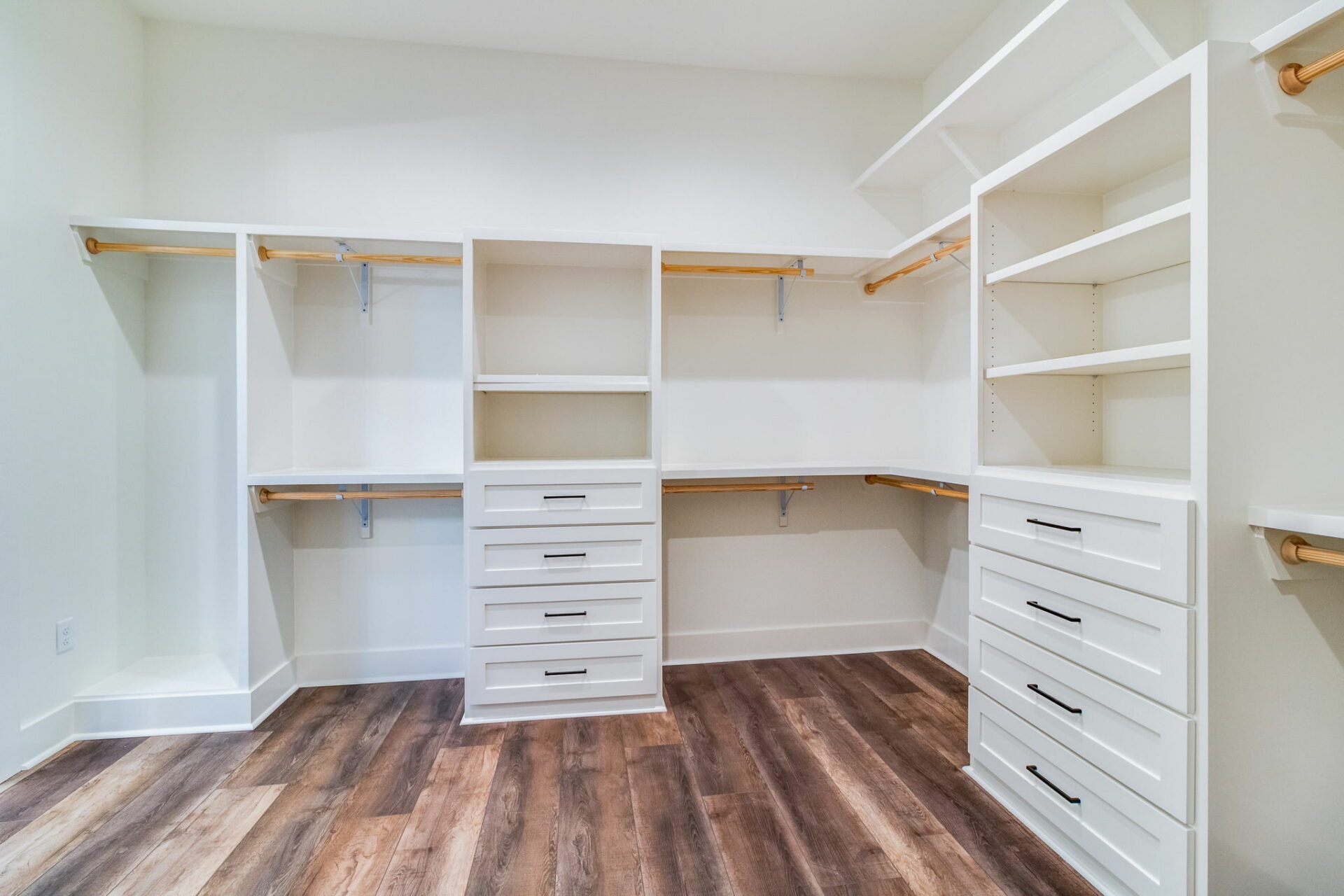 A spacious, empty walk-in closet featuring white Waltman shelving units, drawers, and multiple hanging rods on the walls. The wood floor has a polished finish, and the walls are painted white. The room is well-lit, suggesting ample natural or artificial light.