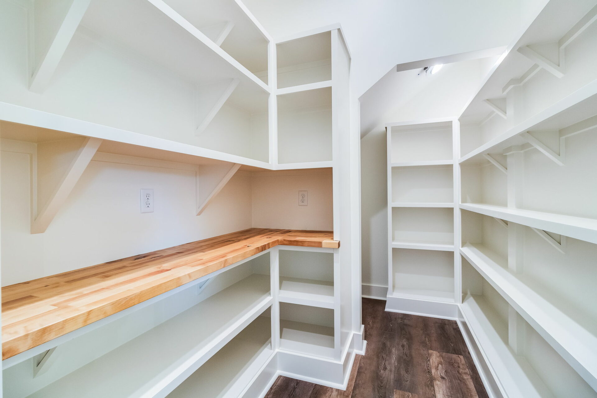 A walk-in pantry with white shelves and a wooden countertop, designed by Waltman. There are multiple storage sections on all sides, offering ample space for organization. The room has a wooden floor and is well-lit with natural and artificial lighting.