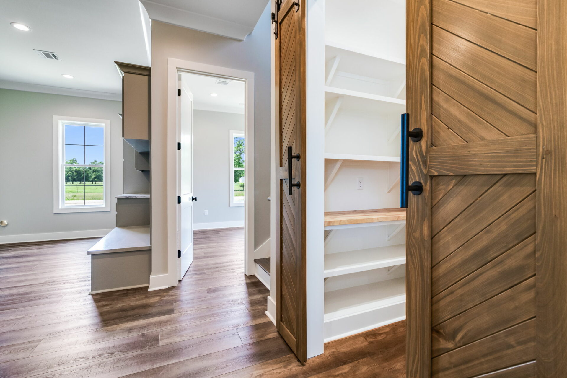 A modern indoor space featuring a Walnut Wood sliding barn door that opens to a white pantry with built-in shelves. Adjacent are a room with large windows, wooden stairs, and another door. The floor is wooden, and the walls are painted in light colors.