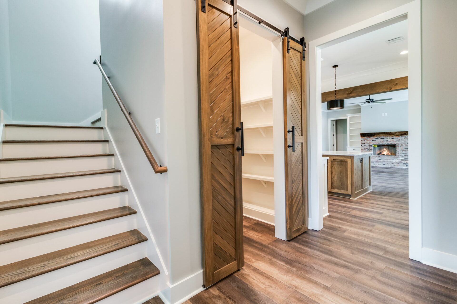 A modern home interior by Waltman features a wooden staircase on the left, wooden sliding barn doors in the center, and an open kitchen and living area with a brick fireplace visible in the background. The flooring is a dark wood laminate.